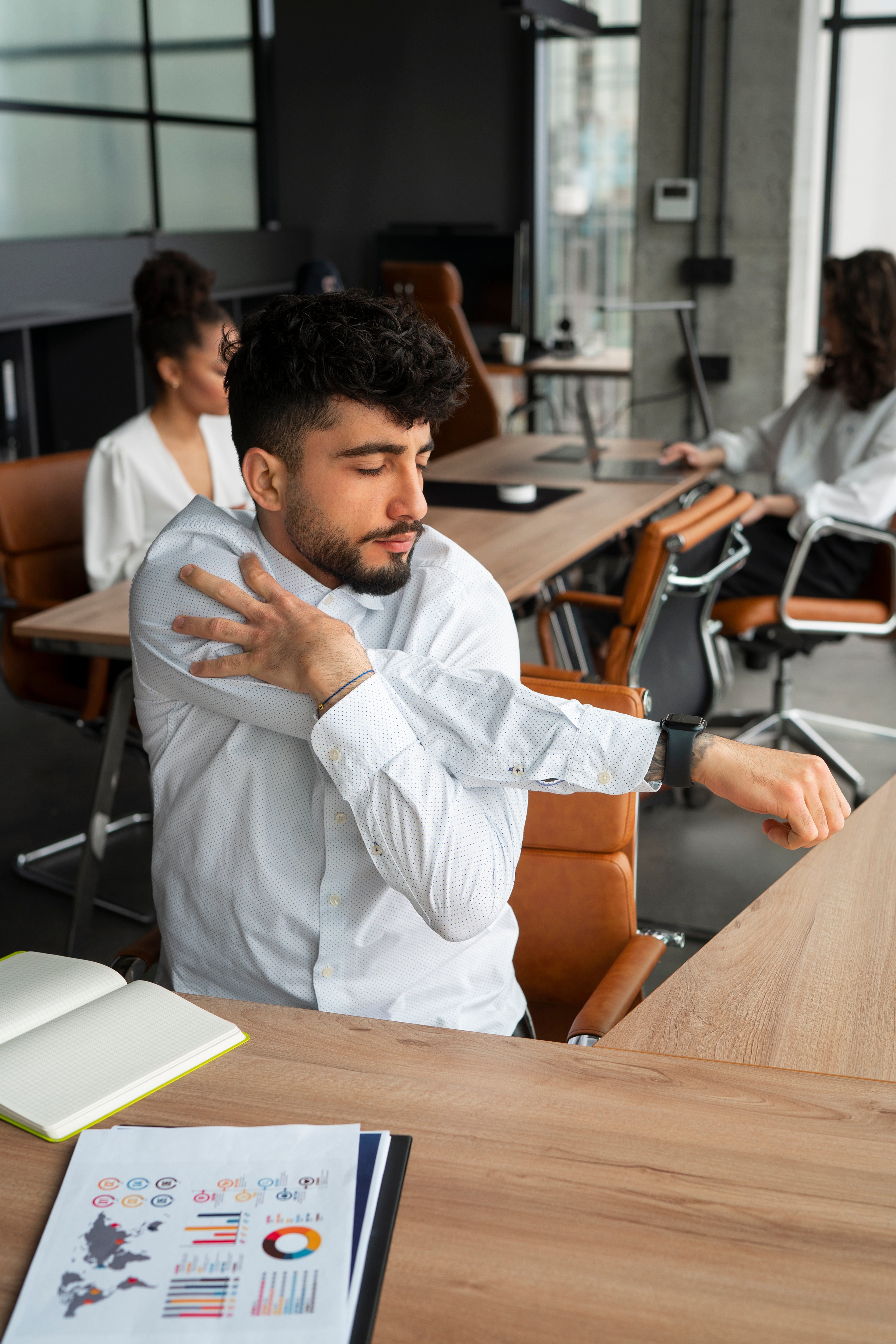 Trabalhador sentado em uma mesa de escritório realiza alongamento do ombro e do braço durante o expediente, em ambiente corporativo moderno, ilustrando a importância de pausas e cuidados ergonômicos no trabalho.