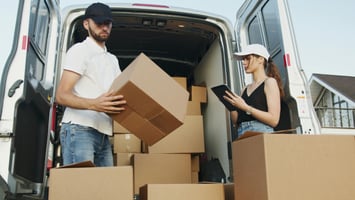 Dois trabalhadores estão descarregando caixas de papelão de uma van branca. Um homem, usando boné preto, camiseta polo branca e jeans, carrega uma caixa grande nos braços. Ao lado dele, uma mulher com boné branco, blusa preta e shorts jeans segura um tablet e parece estar conferindo ou registrando informações sobre as entregas. No fundo, dentro da van, há várias outras caixas empilhadas. A cena acontece em área externa, próxima a uma casa.