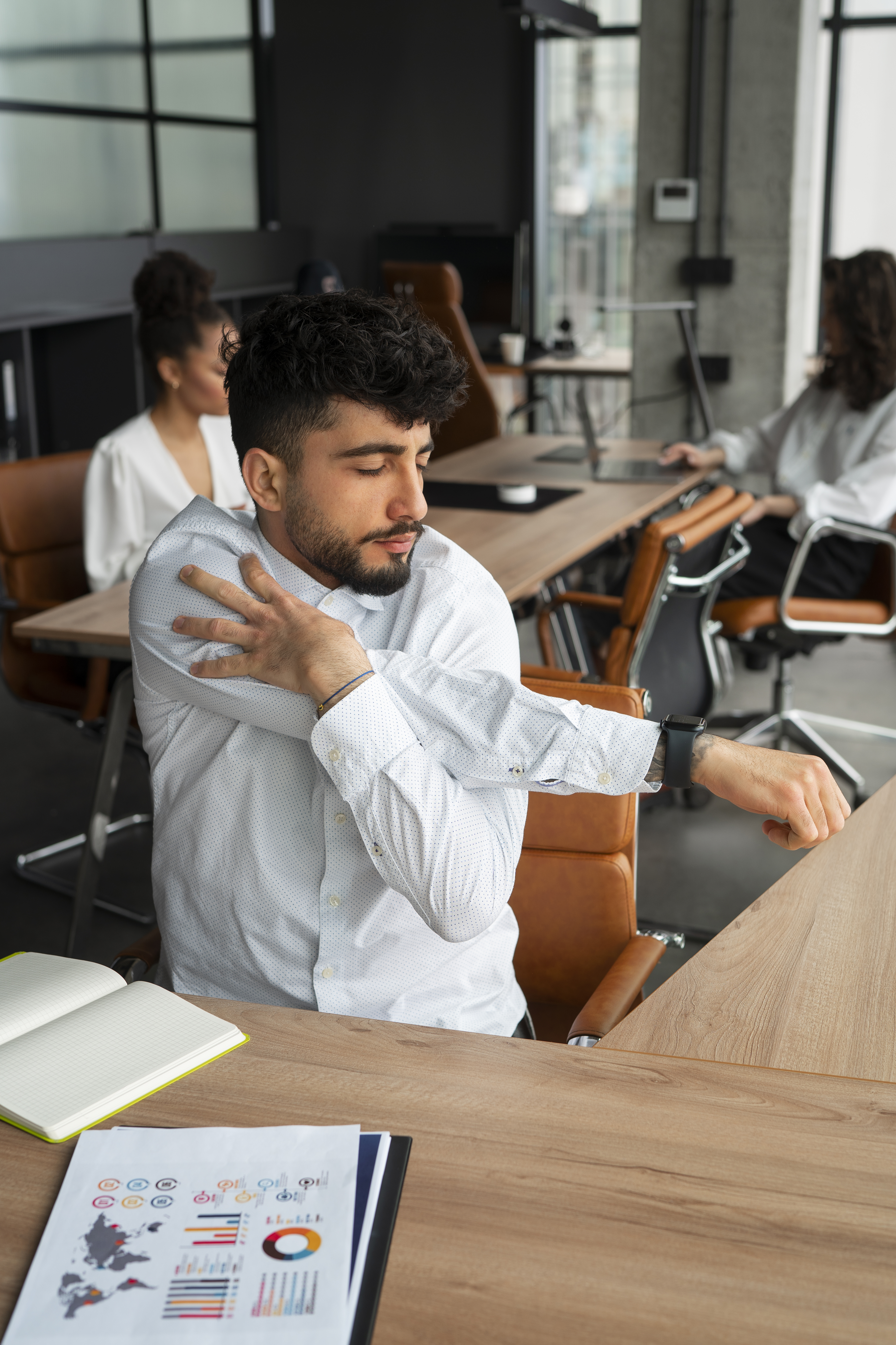 Trabalhador sentado em uma mesa de escritório realiza alongamento do ombro e do braço durante o expediente, em ambiente corporativo moderno, ilustrando a importância de pausas e cuidados ergonômicos no trabalho.