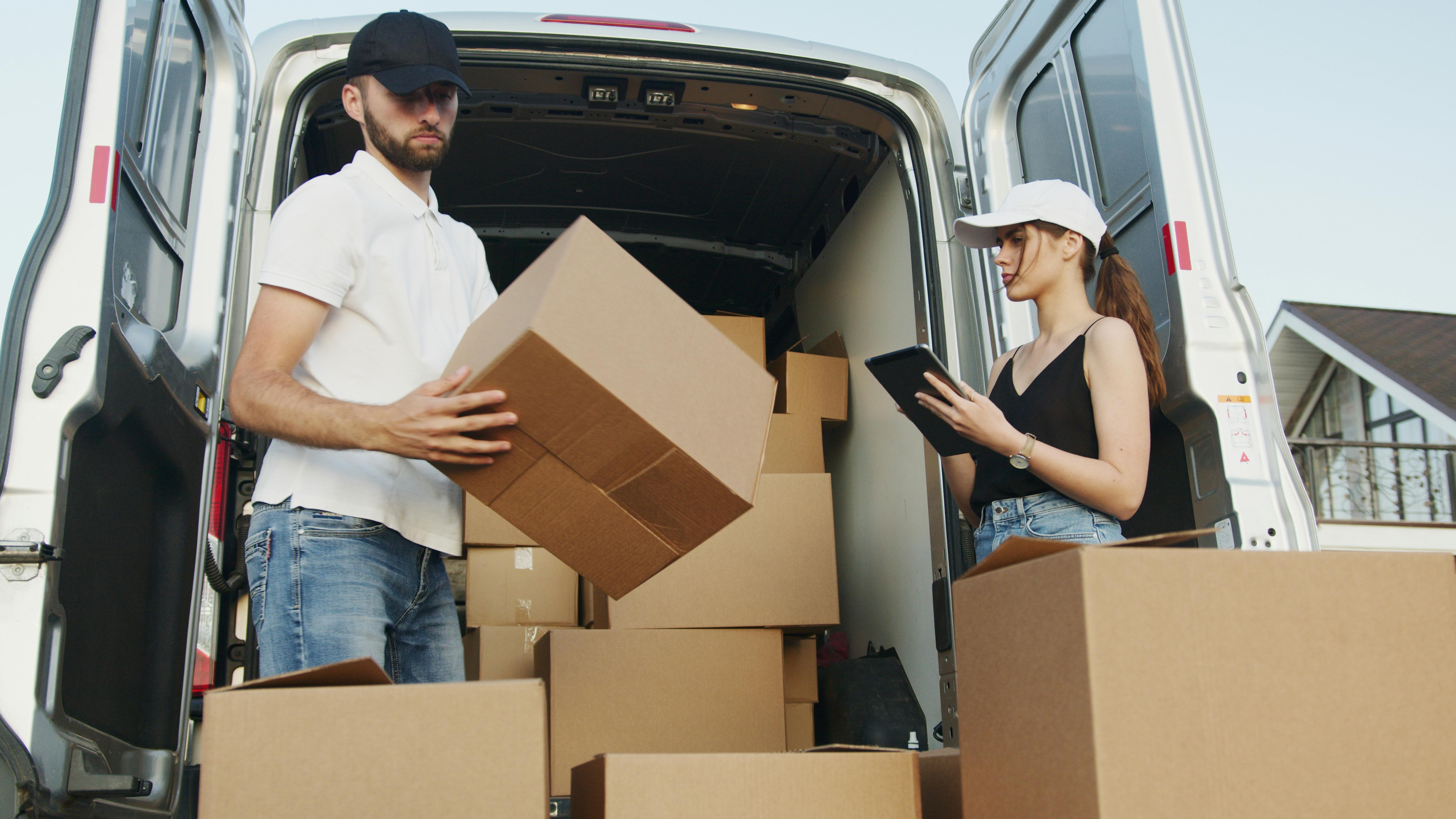 Dois trabalhadores estão descarregando caixas de papelão de uma van branca. Um homem, usando boné preto, camiseta polo branca e jeans, carrega uma caixa grande nos braços. Ao lado dele, uma mulher com boné branco, blusa preta e shorts jeans segura um tablet e parece estar conferindo ou registrando informações sobre as entregas. No fundo, dentro da van, há várias outras caixas empilhadas. A cena acontece em área externa, próxima a uma casa.