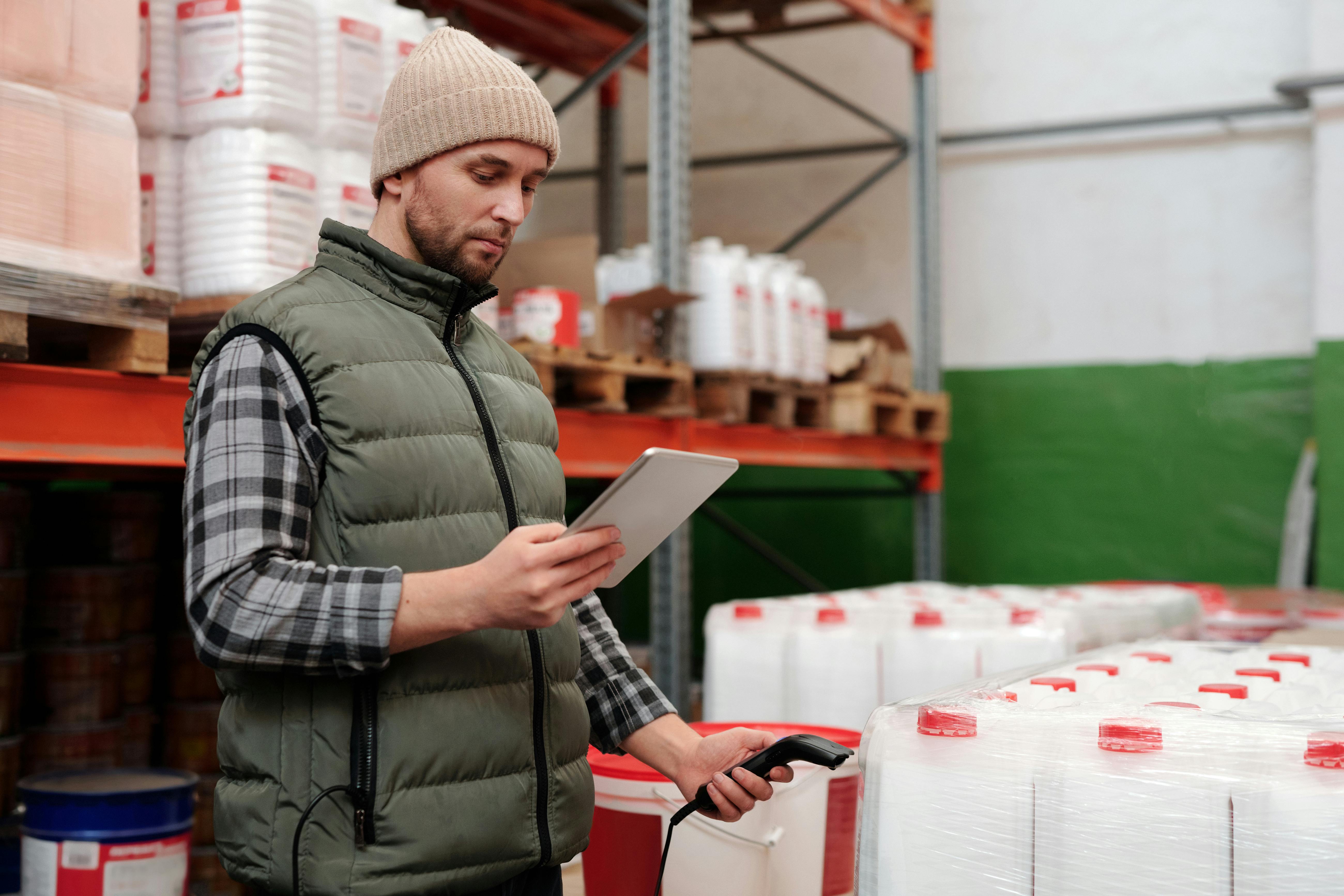 Trabalhador de armazém usando um tablet e um leitor de código de barras para registrar produtos embalados em galões brancos com tampas vermelhas, organizados em palets.